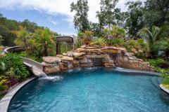 Waterslide framed by vibrant tropical plants and stonework in a custom outdoor pool setting. Waterslide framed by vibrant tropical plants and stonework in a custom outdoor pool setting.