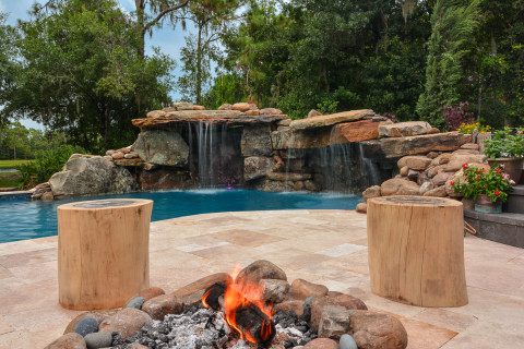Sitting by the poolside fire pit with two log seats and a view of the backyard pool waterfall